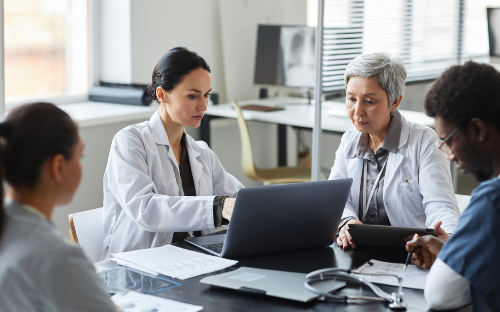 Brunette Female Clinician In Lab Coat Pointing At Laptop Screen